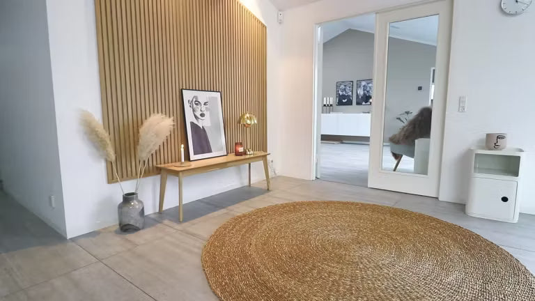 Minimalt nordiskt vardagsrum med en rund jute-matta, träbänk med pampasgräs, inramad konst och en accentvägg i Akupanel akustiskt trä med lameller.