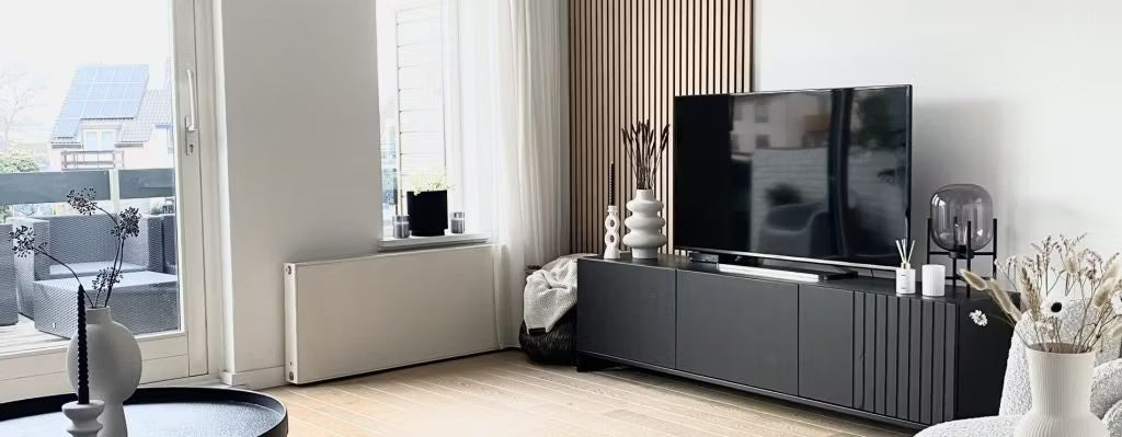 Modernt vardagsrum med svart TV-möbel, platt-TV, dekorativa vaser och stora fönster med naturligt ljusinsläpp mot uteplatsen; Akupanel på ena väggen.