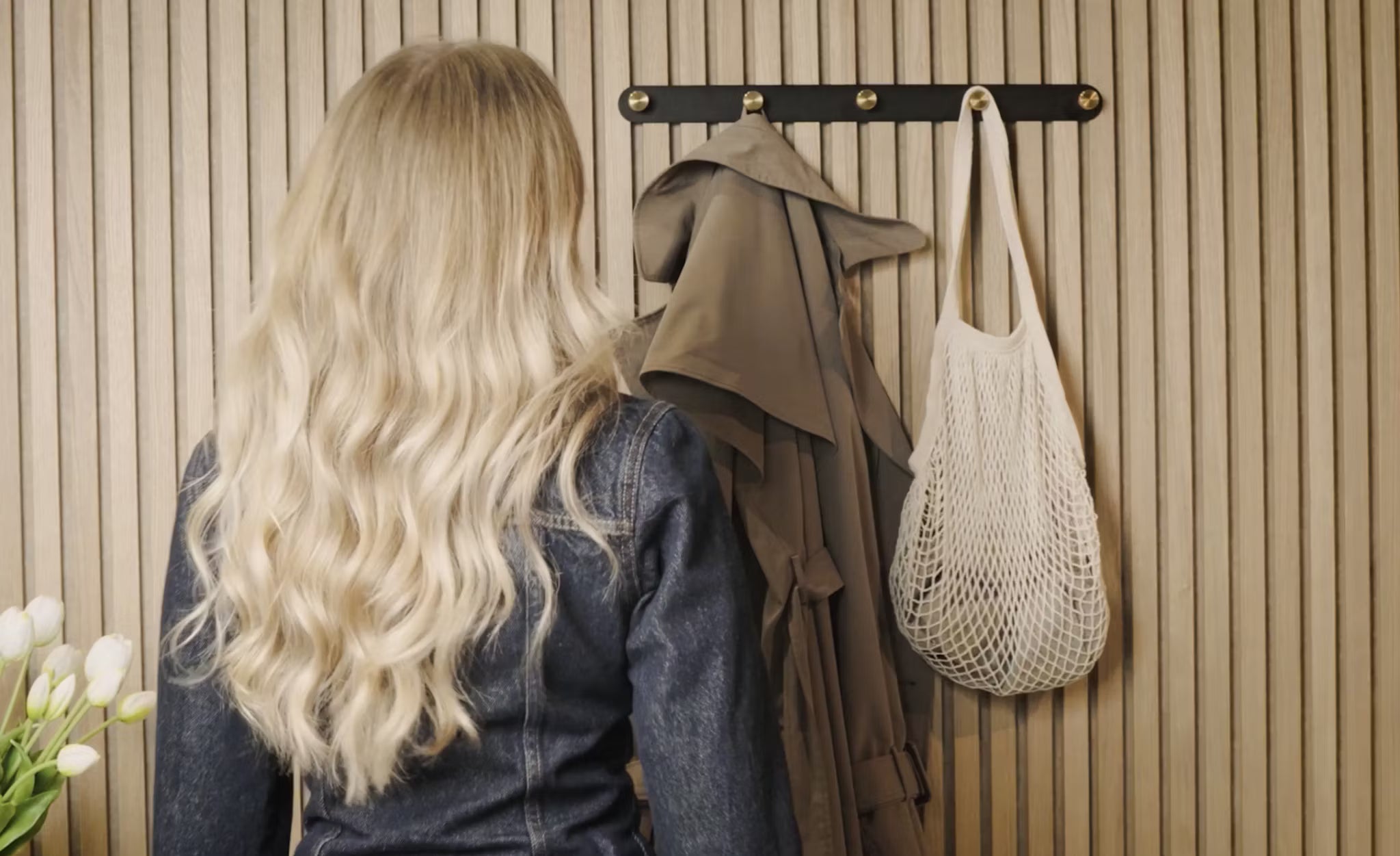 En kvinna med långt blont hår står framför en klädhängare med en trenchcoat, en vit nätväska och färska vita tulpaner bredvid en akustisk Akupanel-träpanel i hörnet.