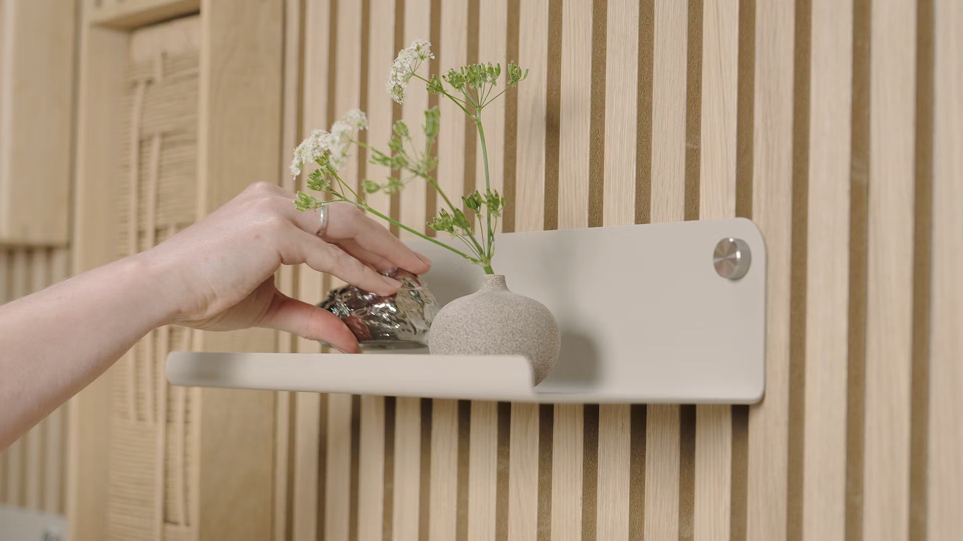 En hand placerar ett litet dekorativt föremål bredvid en vas med vita blommor på en minimalistisk hylla mot en strukturerad nordisk akustisk träpanel.