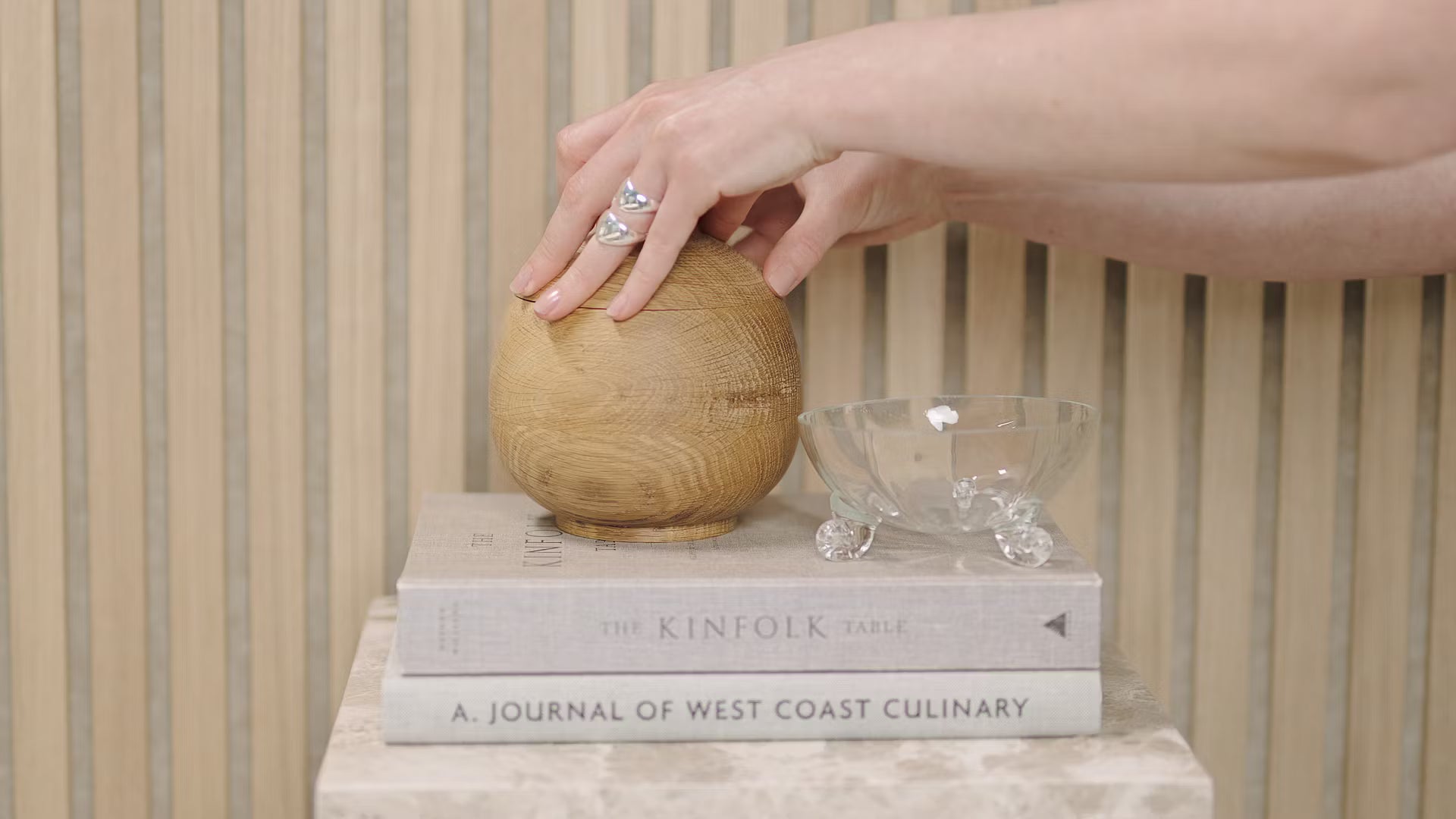 En hand rör vid en träkula som är placerad på staplade böcker, däribland Kinfolk Table och Journal of West Coast Culinary, bredvid en glasskål på en akustisk panel av träribbor.