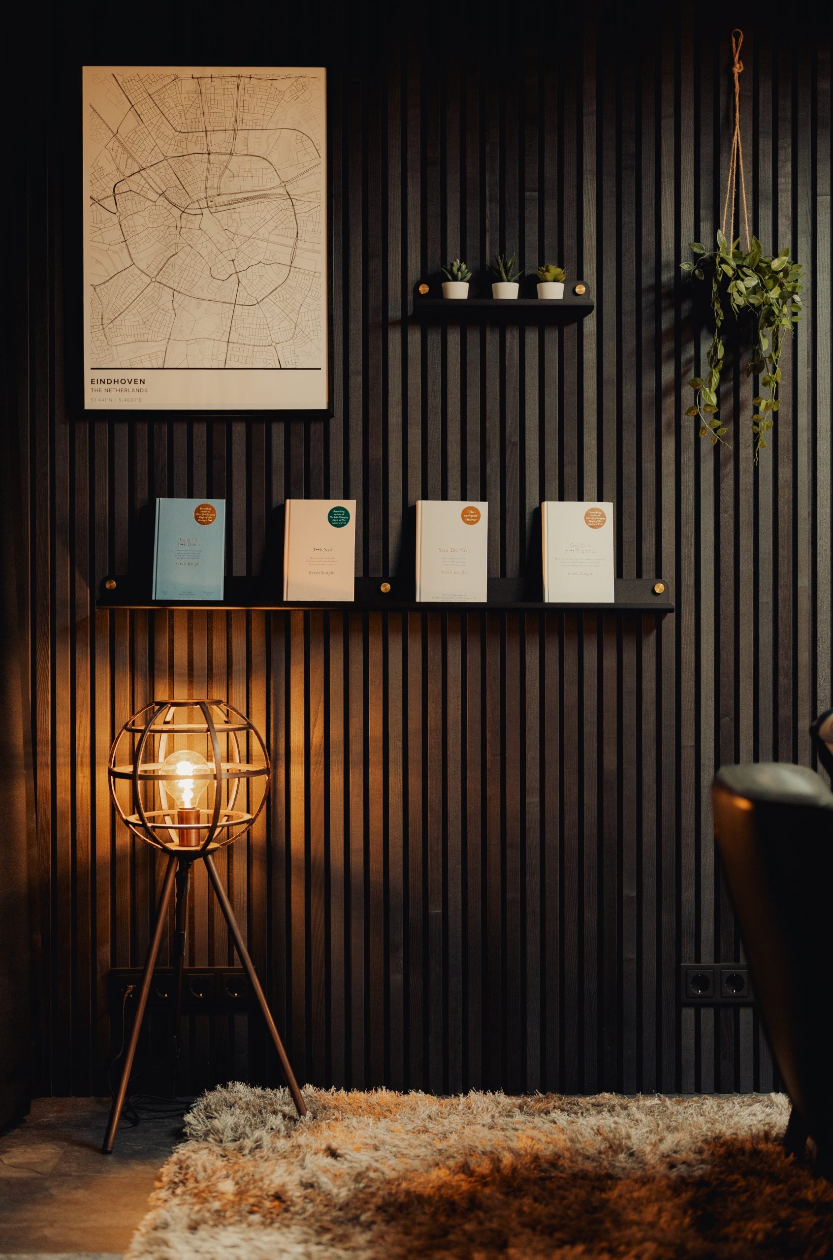 Modern Nordic room with glowing lamp, shag rug, and wall shelves holding books, small plants, and an acoustic panel featuring a framed travel map above.