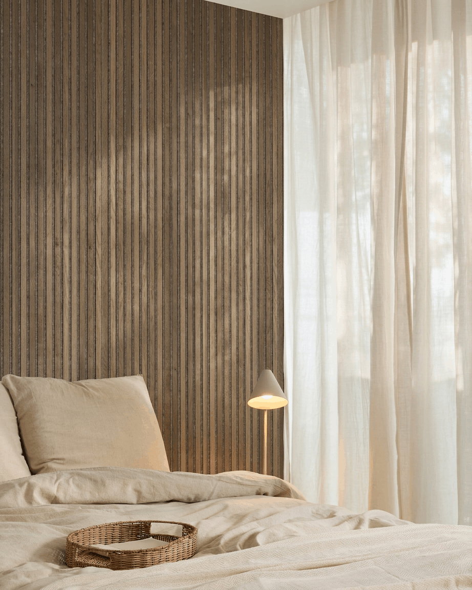 Minimalistiskt sovrum med beige sängkläder, en bricka i rotting, WoodUpp Akupanel Rustic Grey Oak akustisk ribbväggspanel, en vit lampa och skira gardiner.