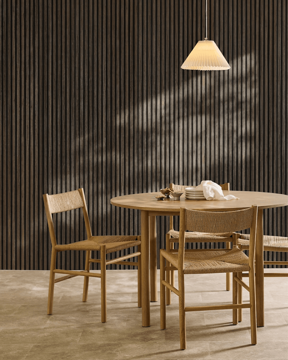 Ett runt bord med fyra flätade stolar står bredvid en WoodUpp Akupanel Black Oak Akustikpanel, under en veckad pendellampa.