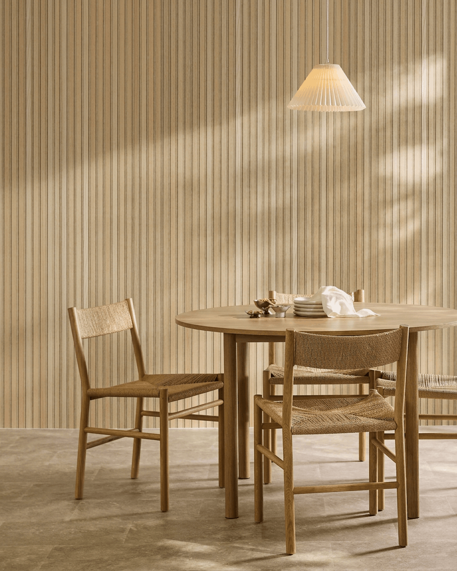 Minimalistisk matplats med runt bord, vävda stolar och pendellampa intill en WoodUpp Akupanel akustikpanel i trä i Classic Oak med beige filt.