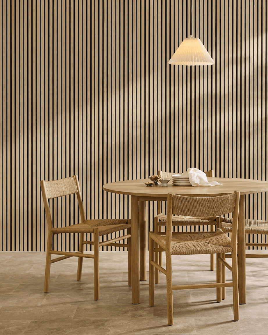 Minimalistisk matplats med ett runt bord, flätade stolar, pendellampa och en WoodUpp Akupanel Classic Oak Akustikpanel monterad på väggen.