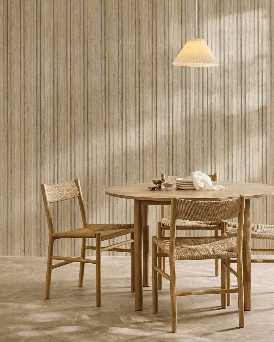 Minimalistisk matplats med ett runt träbord, flätade stolar, en pendellampa och en WoodUpp Akupanel akustikpanel i trä i Classic Oak med grey filt.