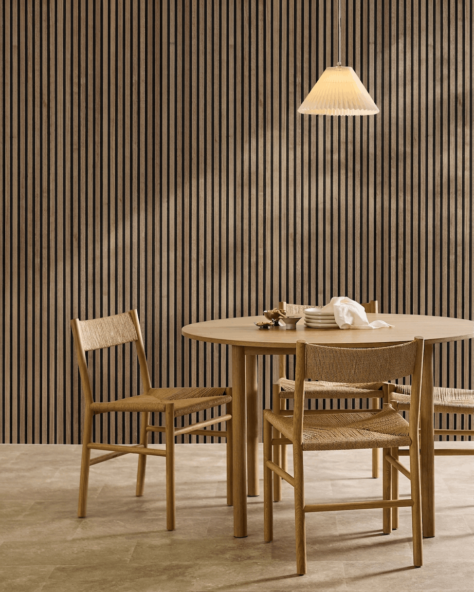 Minimalistisk matplats med runt bord, flätade stolar, pendellampa och en WoodUpp Akupanel akustikpanel i trä i Natural Wood med black filt på väggen.