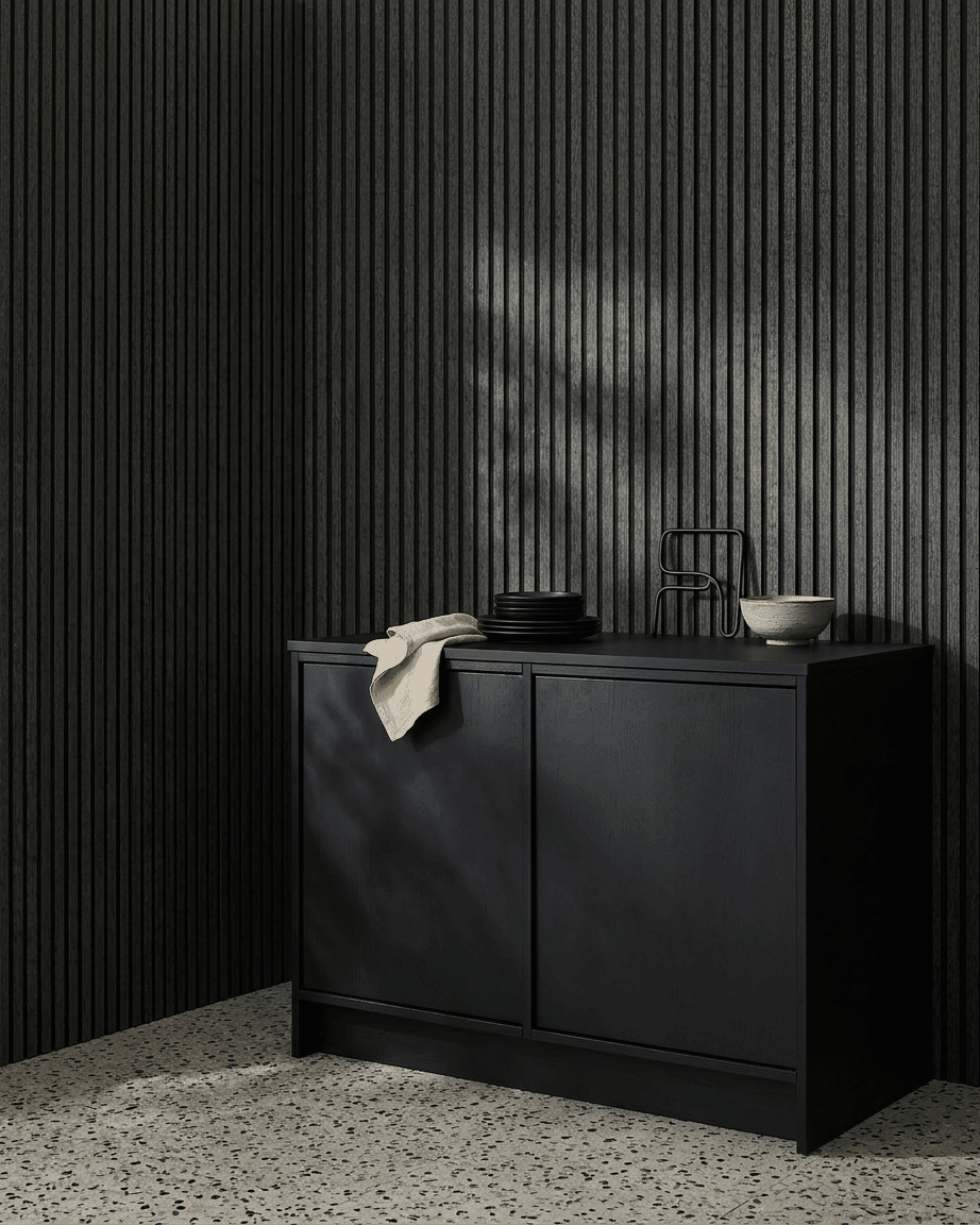 Ett black skåp bredvid en WoodUpp Akupanel akustikpanel i trä i Charcoal/Black Felt, med skålar och tallrikar ovanpå, på ett ljust terrazzogolv.