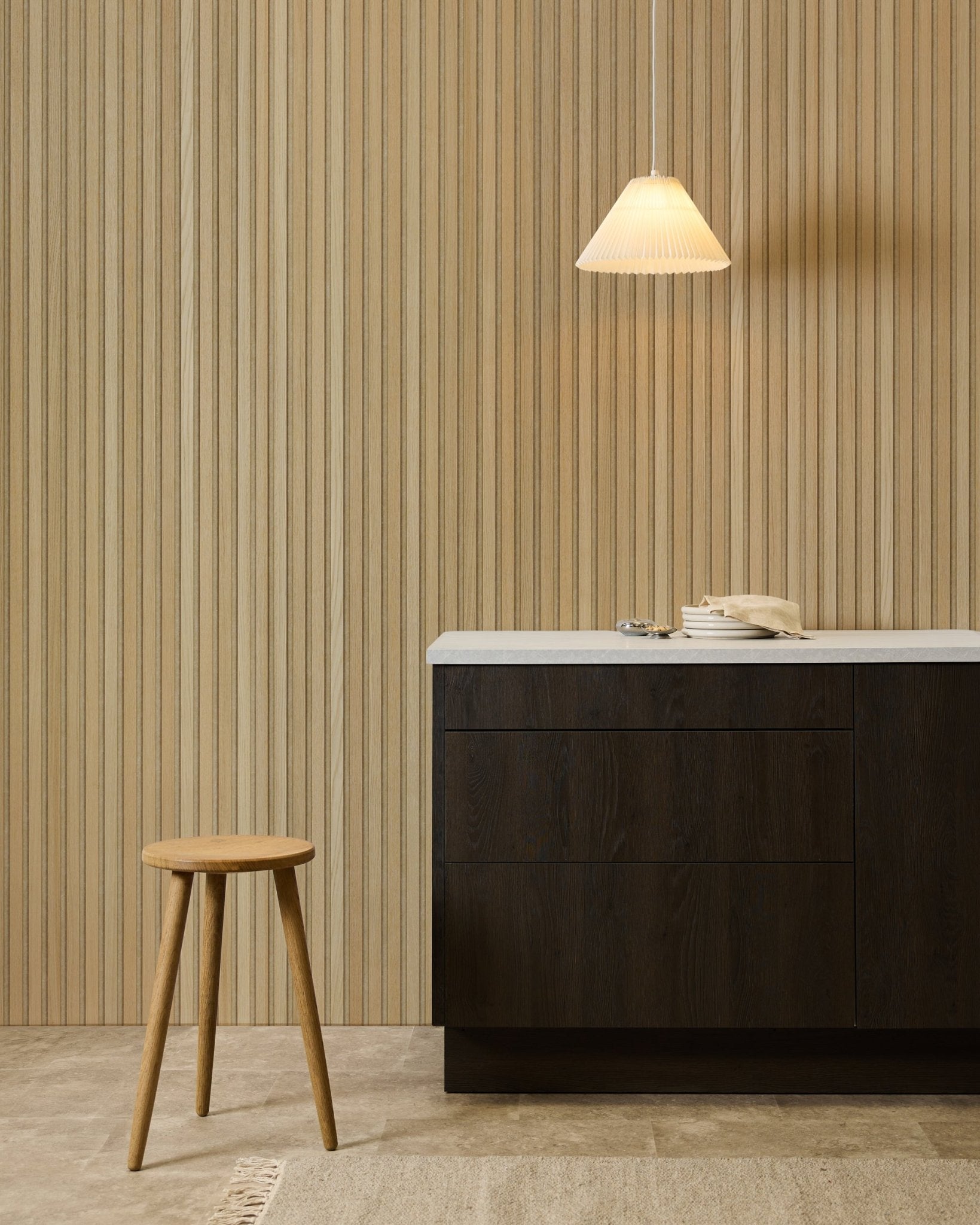 Minimalist kitchen with WoodUpp Akupanel Classic Oak acoustic panel on the wall, wooden stool, dark cabinet, beige rug, and rotary phone on a white countertop.