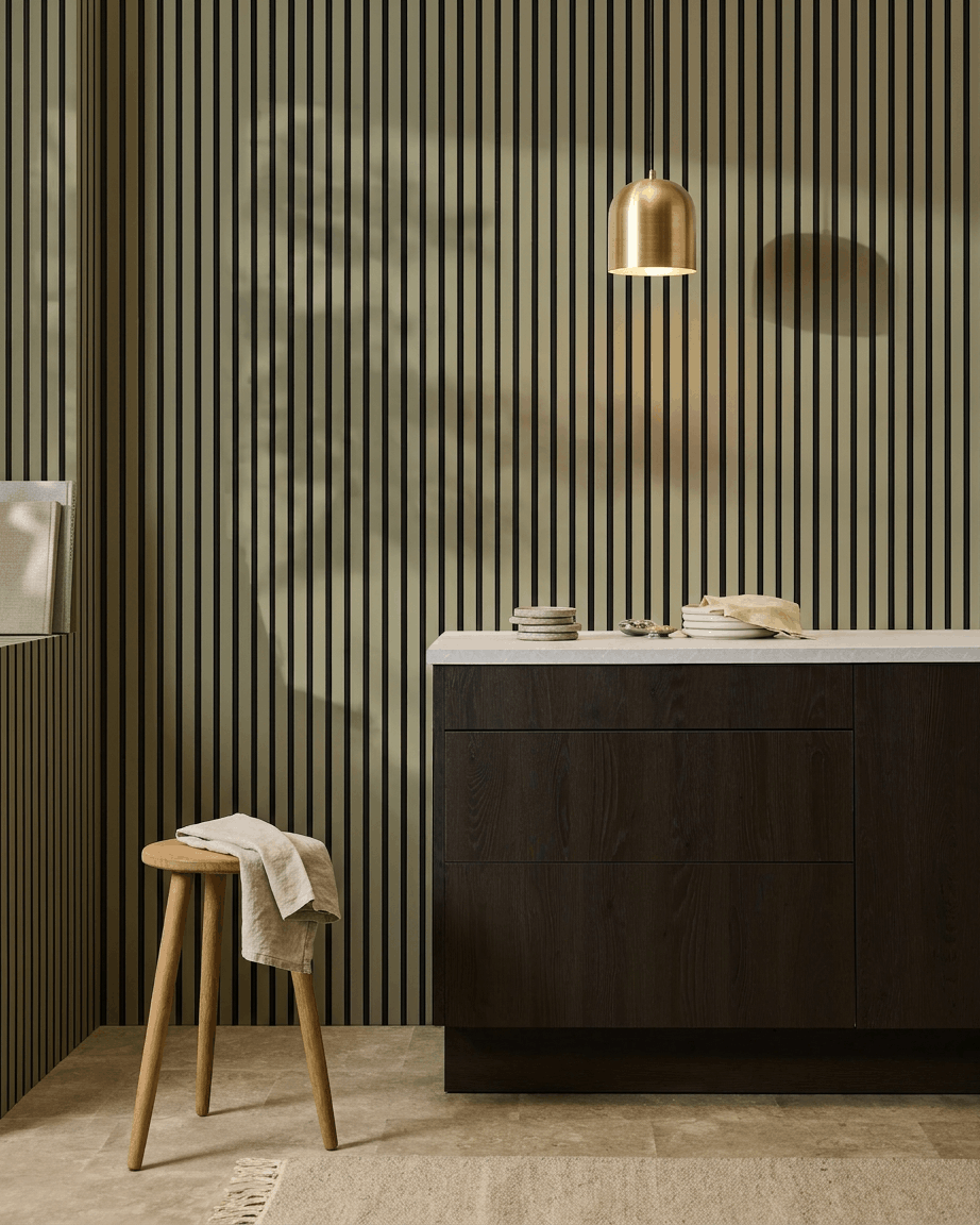 Modernt kök med mörka träskåp, bänkskiva i marmor, brass pendellampa och en WoodUpp Akupanel Colored akustisk slatwall-panel för ljudabsorption.