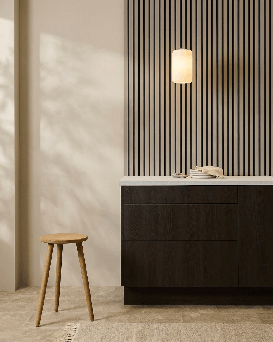 Minimalistiskt kök med WoodUpp Akupanel (Stone, Black filt) akustikpaneler på väggen, träpall, mörka skåp och pendellampa i neutrala toner.