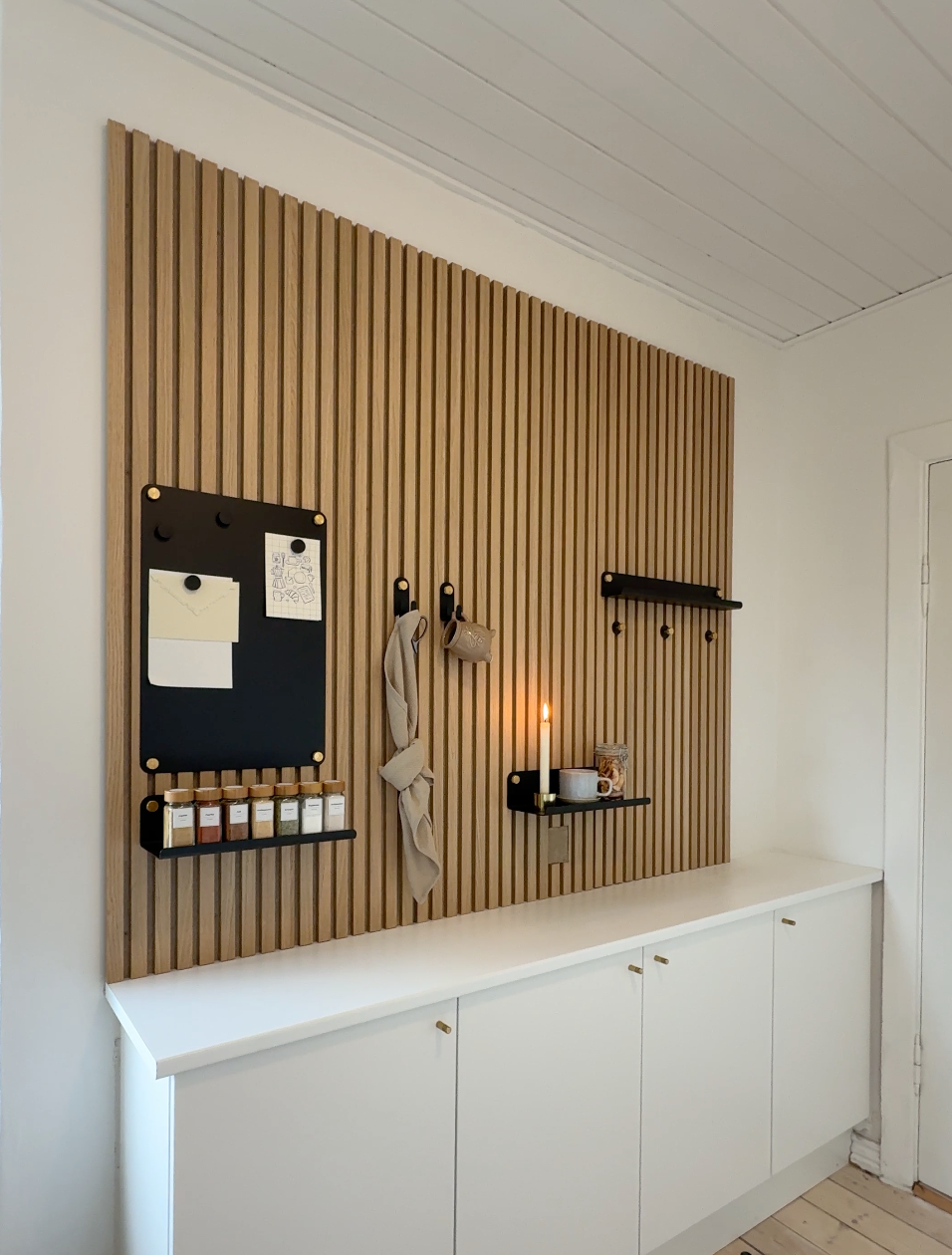 Minimalist entryway with a vertical Akupanel acoustic wood slat wall, black hooks, shelves, organizer, candle, scarf, and white cabinets on a white wall.