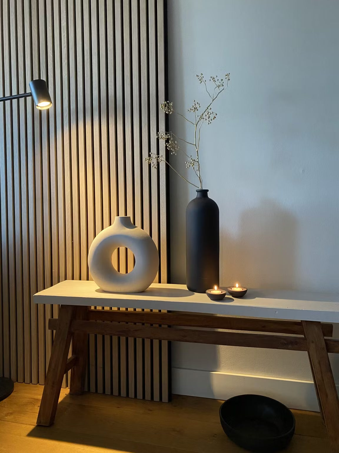 Minimalistisk inredning med en vit cirkulär vas, en svart hög vas med torkade stjälkar, två värmeljus och en svart skål mot en akustisk Akupanel-panel med träribbor.