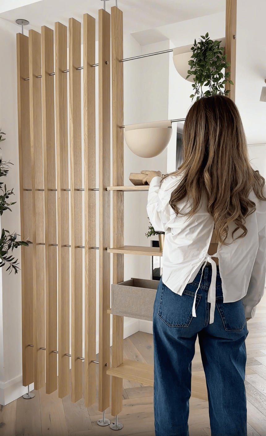 A woman with long hair, wearing a white top and jeans, stands next to a Nordic-style EMBRACE wooden room divider with shelves, greenery, and potted plants.
