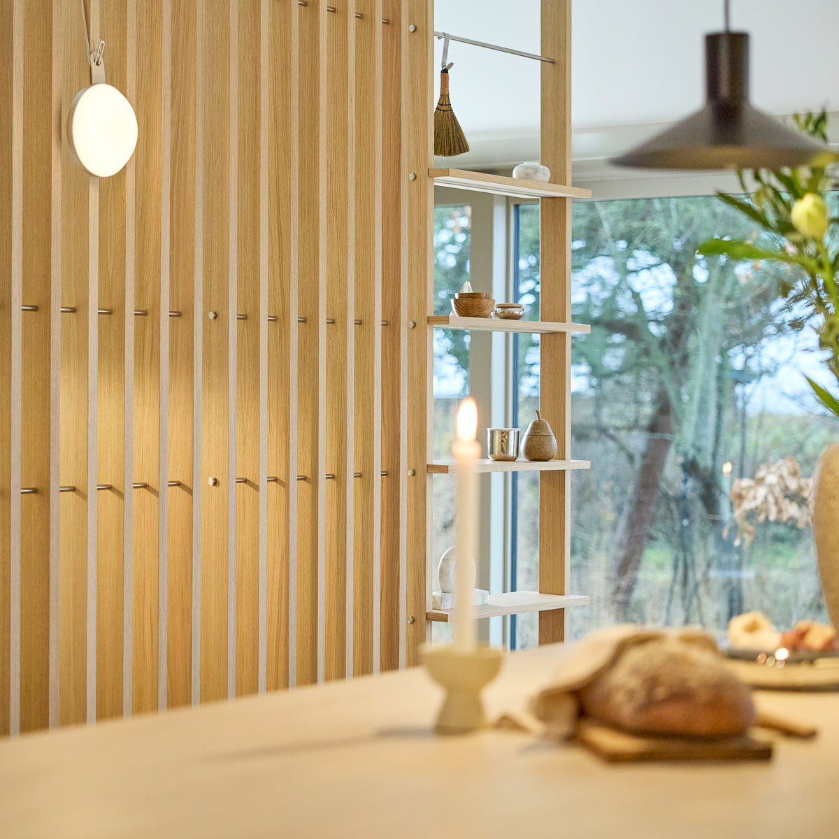 Minimalistiskt interiör med en Nordic EMBRACE rumsavdelare i trä, tänt ljus, färskt bröd på bordet, noga utvald inredning på hyllorna och ett fönster som ramar in träd utanför.