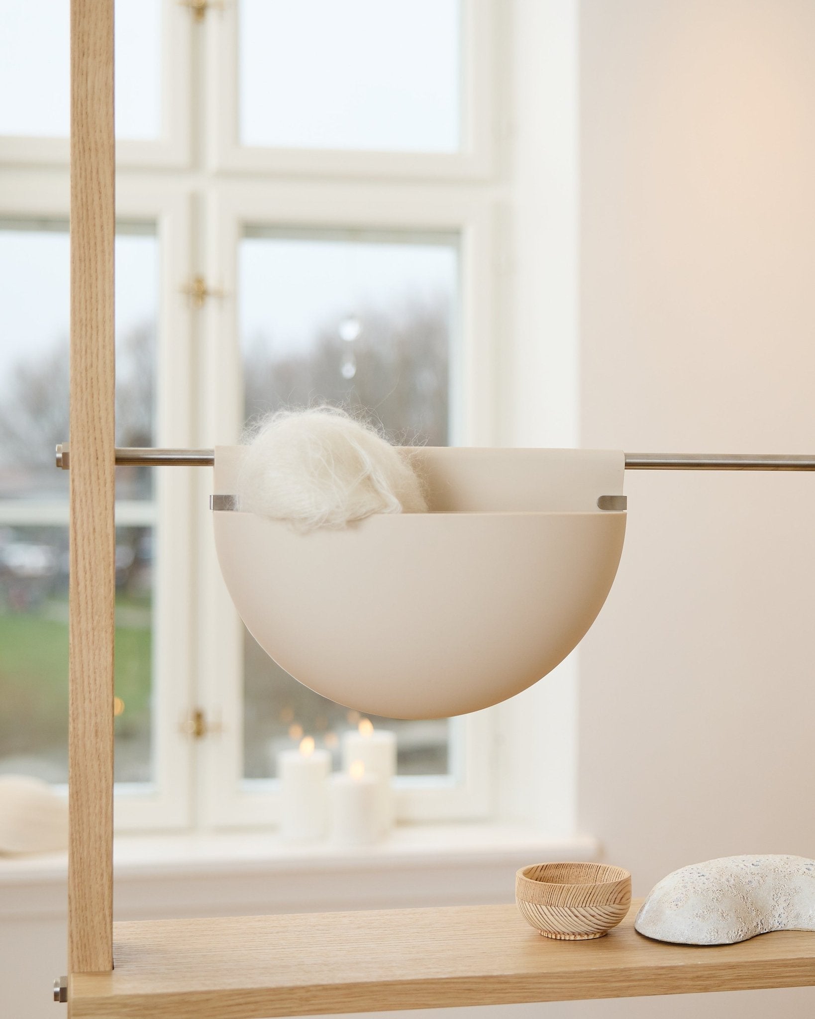En vit skål med ett fluffigt föremål står på en trähylla nära ljus, inredningsdetaljer och akustiktillbehöret WoodUpp Gro Soft Pocket BM2 i ljus beige vid fönstret.