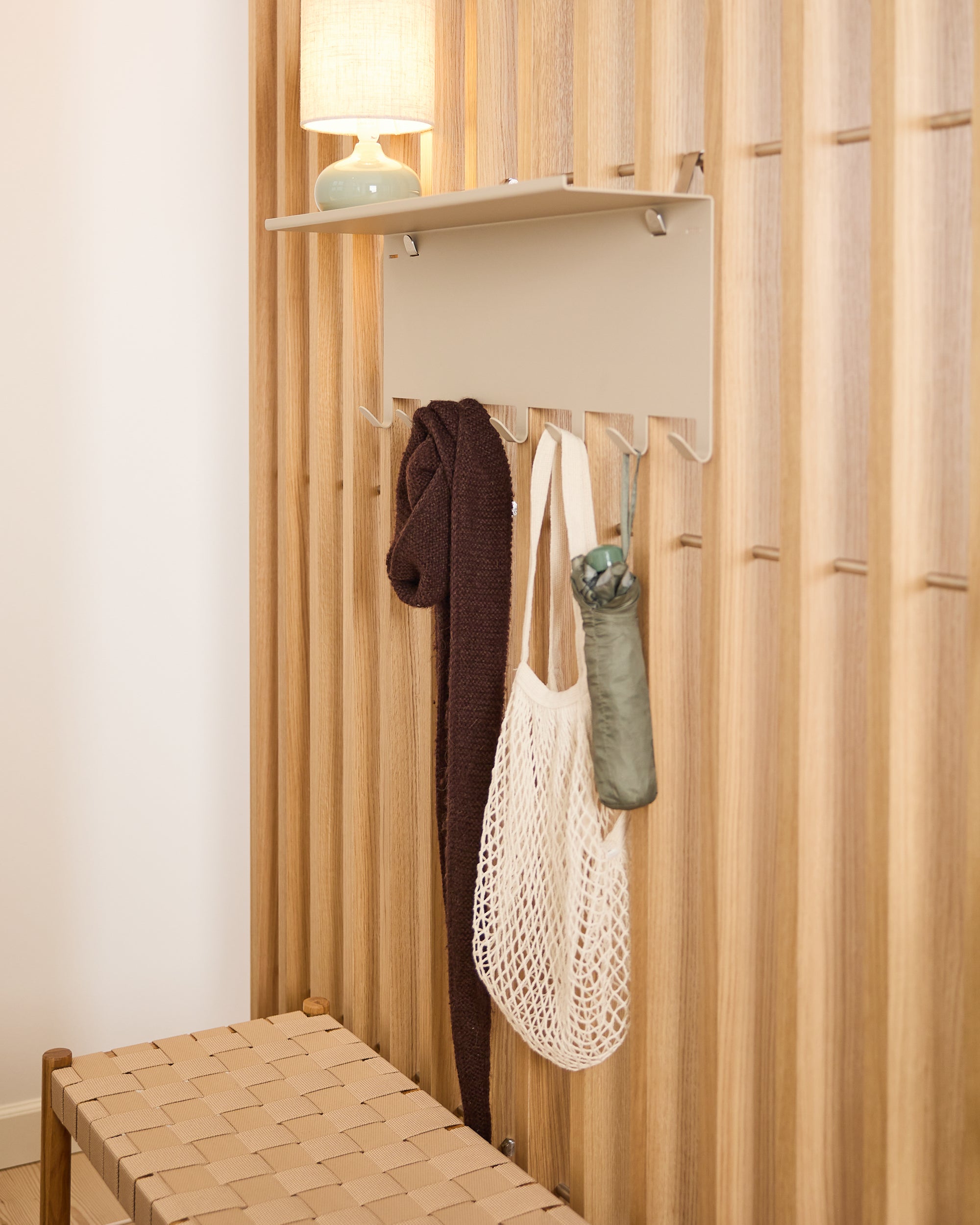 A Nordic-style acoustic slatwall panel with hooks holds a brown scarf, mesh bag, and umbrella; a woven bench sits below a shelf with a lamp.