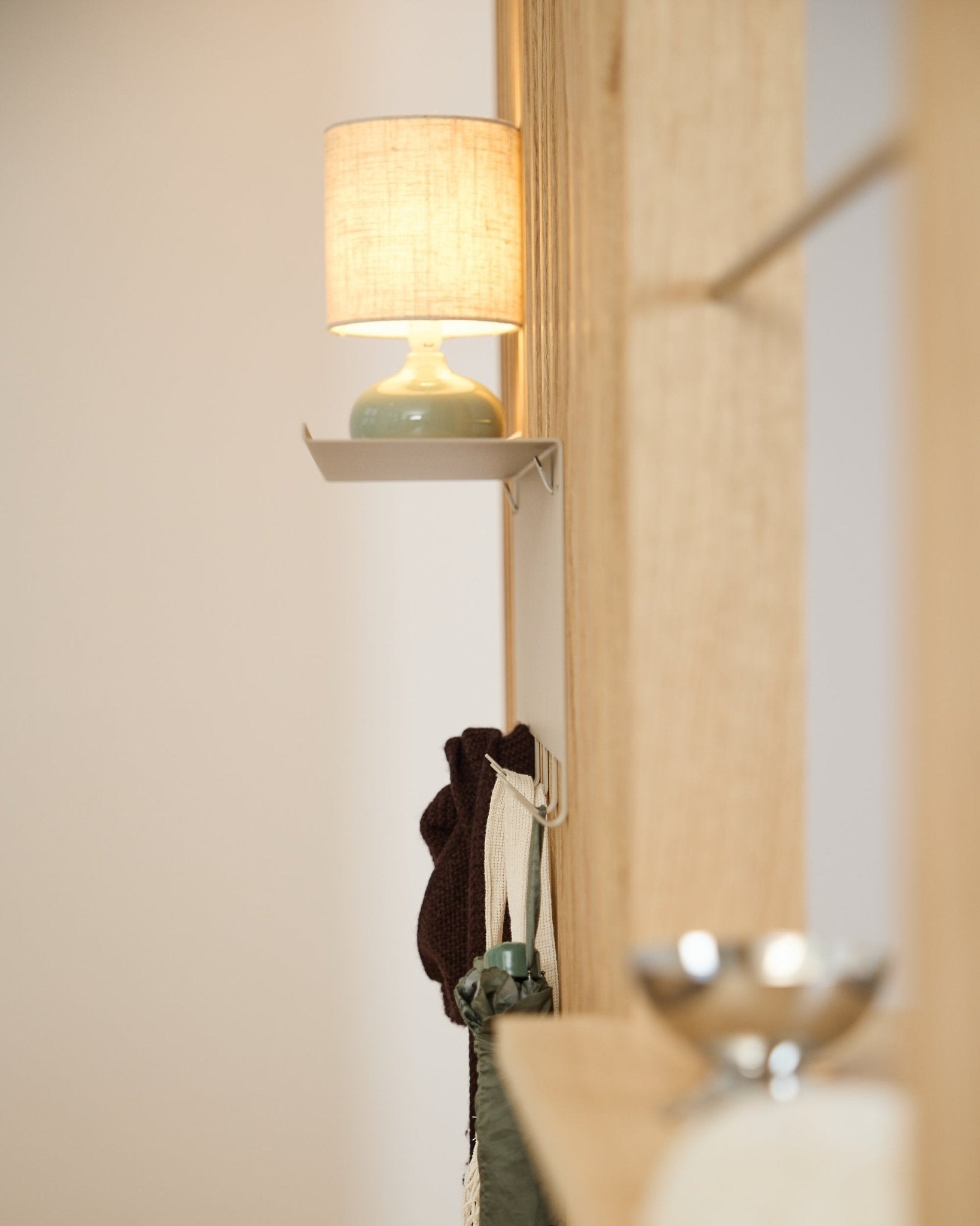 En beige WoodUpp Nell klädhängare BM1 (63,5 x 24 cm) står under en small bordslampa intill en vägg i ljust nordiskt trä Akustikpanel.