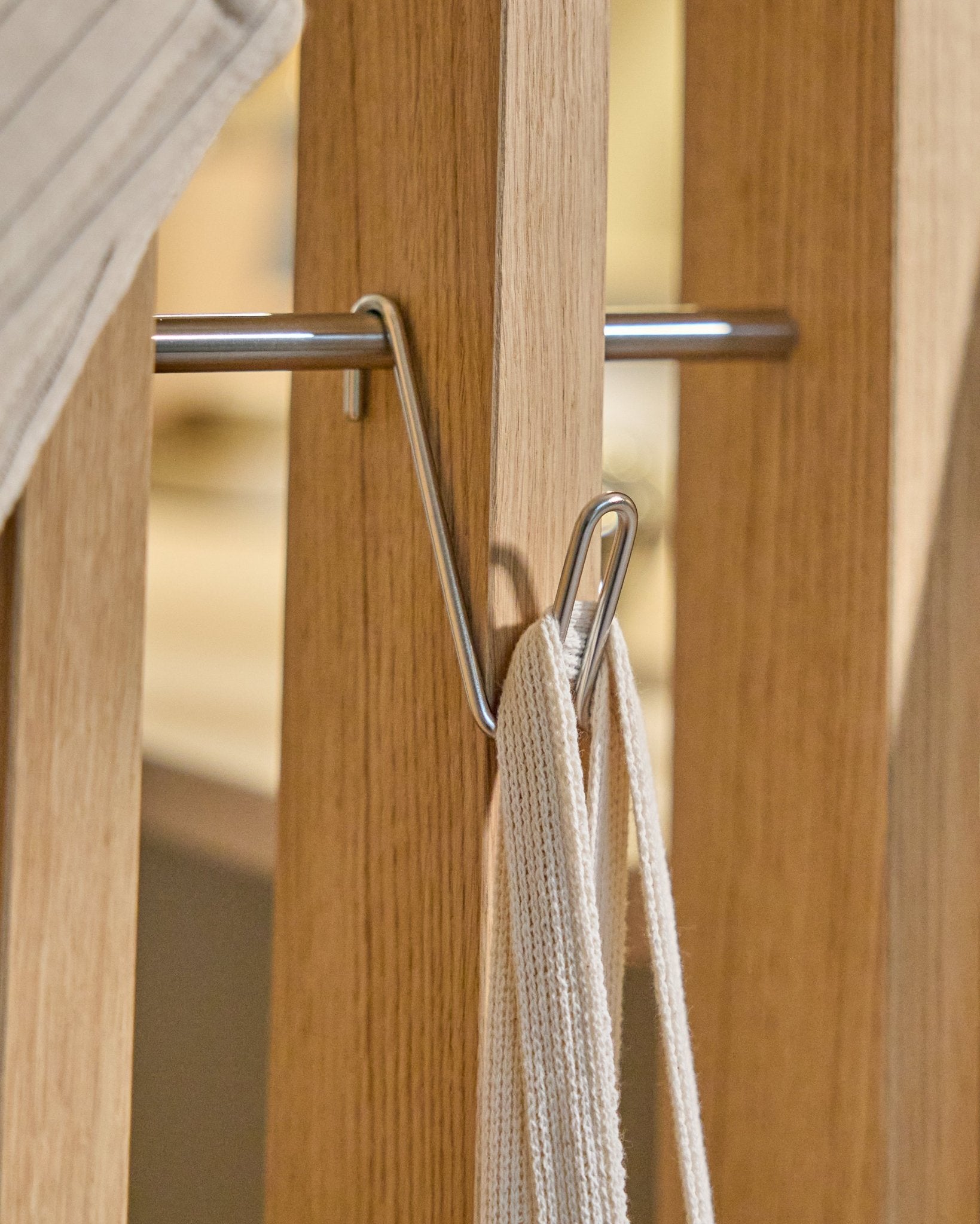 Alva Wire Hook / BM1 (rostfritt Steel, kort) från WoodUpp är monterad på en Akustikpanel skena i trä och håller en vit rem med metallstav mot en beige bakgrund.