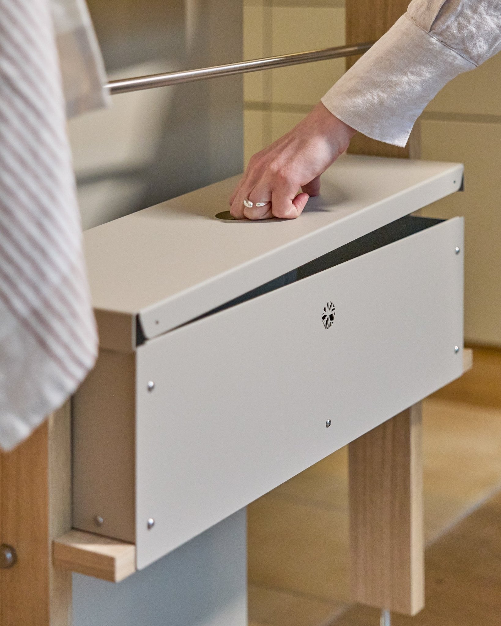 En hand lyfter locket på WoodUpp Nell Metal Box BM2 i beige, med pulverlackerad steel och stöd i trä, formgiven för organiserad nordisk förvaring.