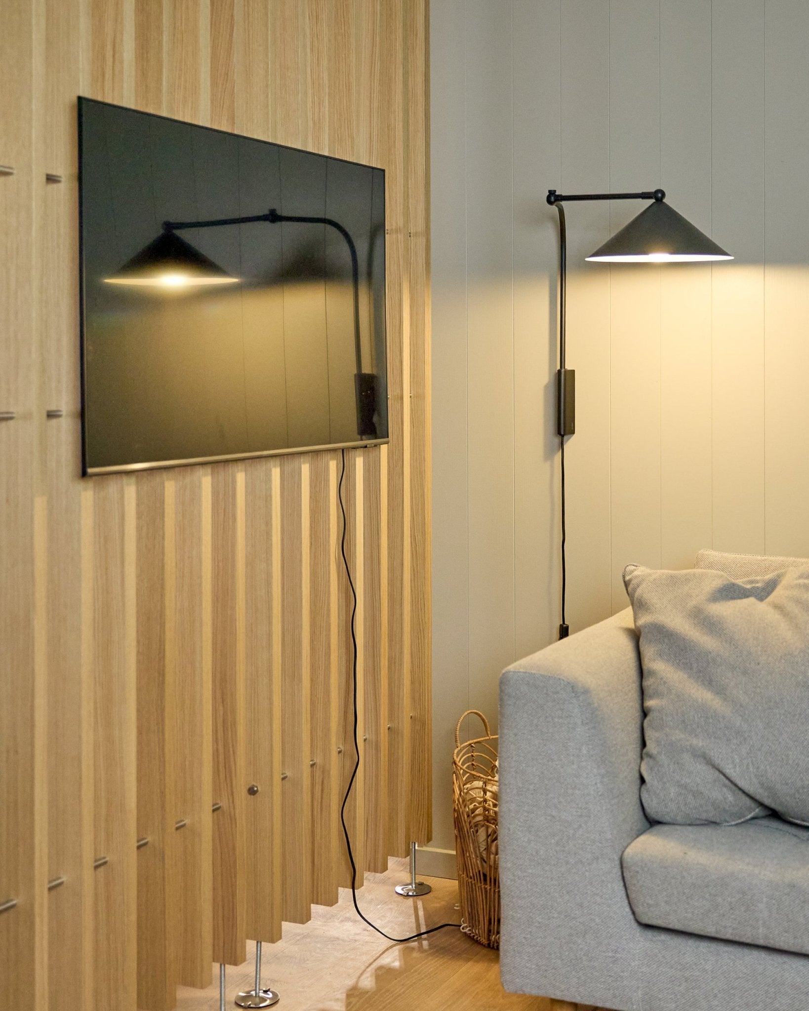 En Nell | TV-Mount / BM1 från WoodUpp (Beige, 64x44 cm) håller en platt-TV på en nordisk Akustikpanel intill en grå soffa; en korg står i närheten.