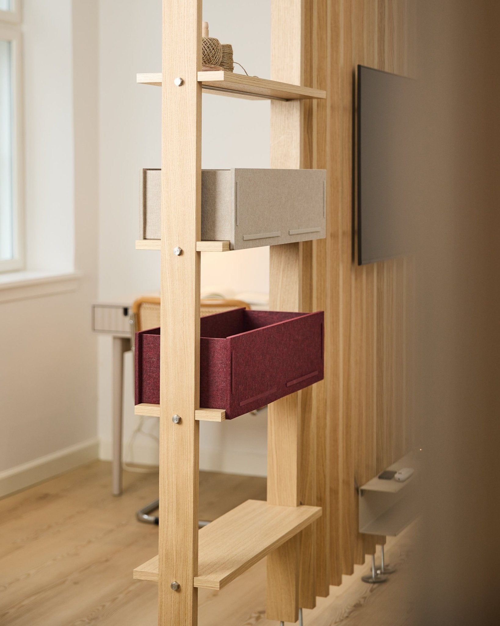En hylla från WoodUpp visar en beige förvaringslåda och en ox red Vigg Felt Box / BM2 i ett ljust, minimalistiskt nordiskt rum med akustiska trädetaljer.
