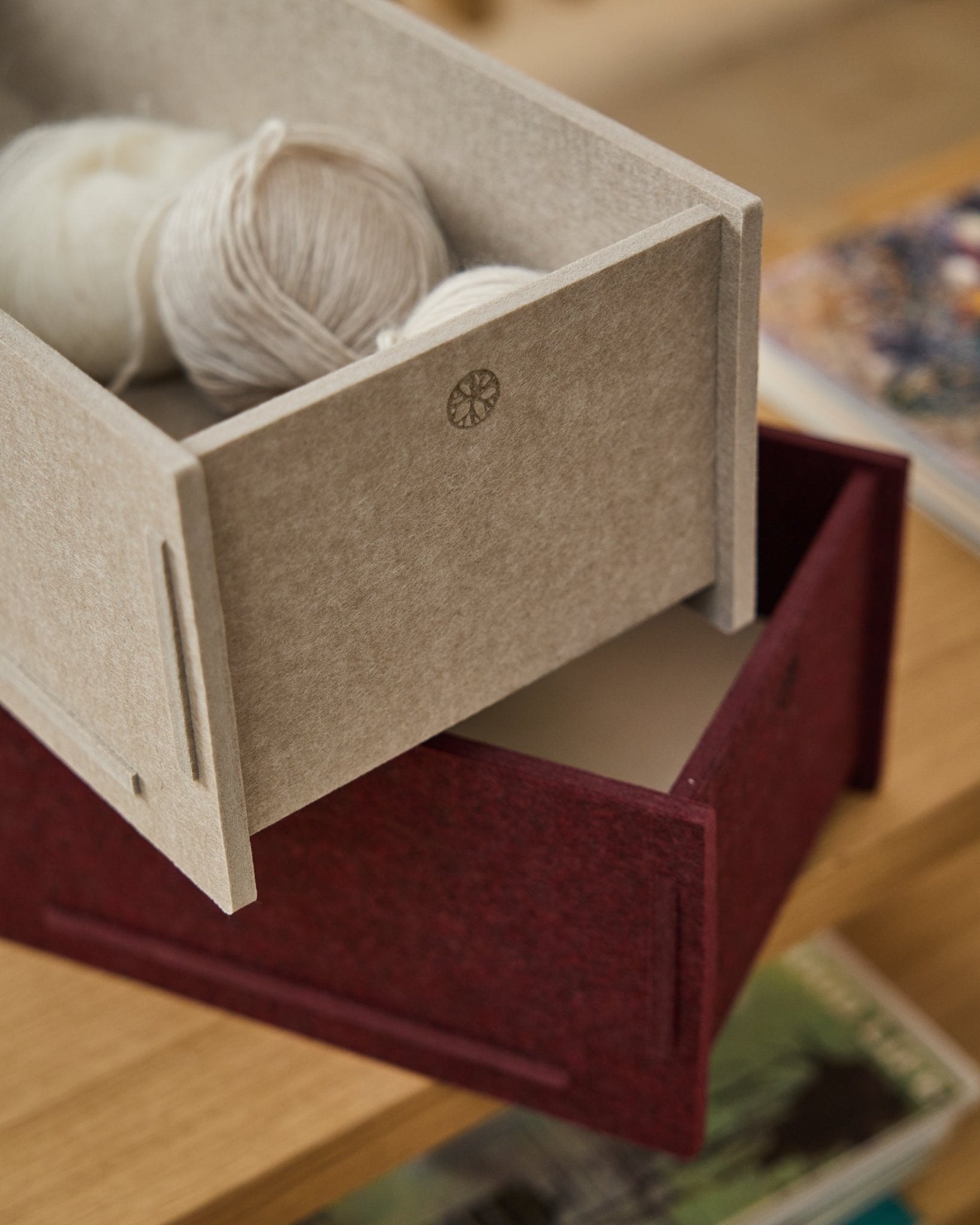 En beige PET-förvaringslåda fylld med garn står ovanpå en stängd WoodUpp Vigg Felt Box/BM2 i ox red, placerad på trägolv med böcker i bakgrunden.