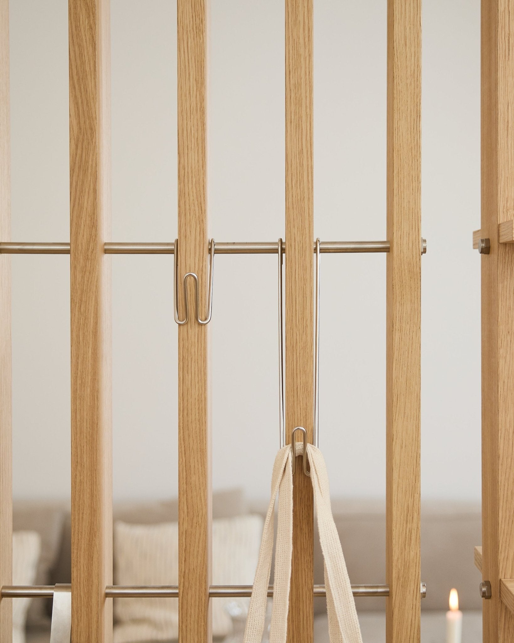 Närbild på WoodUpp Alva | Wire Hook BM1 (rostfritt steel, lång, 3-pack) fäst på ljusa Akustikpanel ribbor med ett vitt band; soffa och ljus är suddiga i bakgrunden.