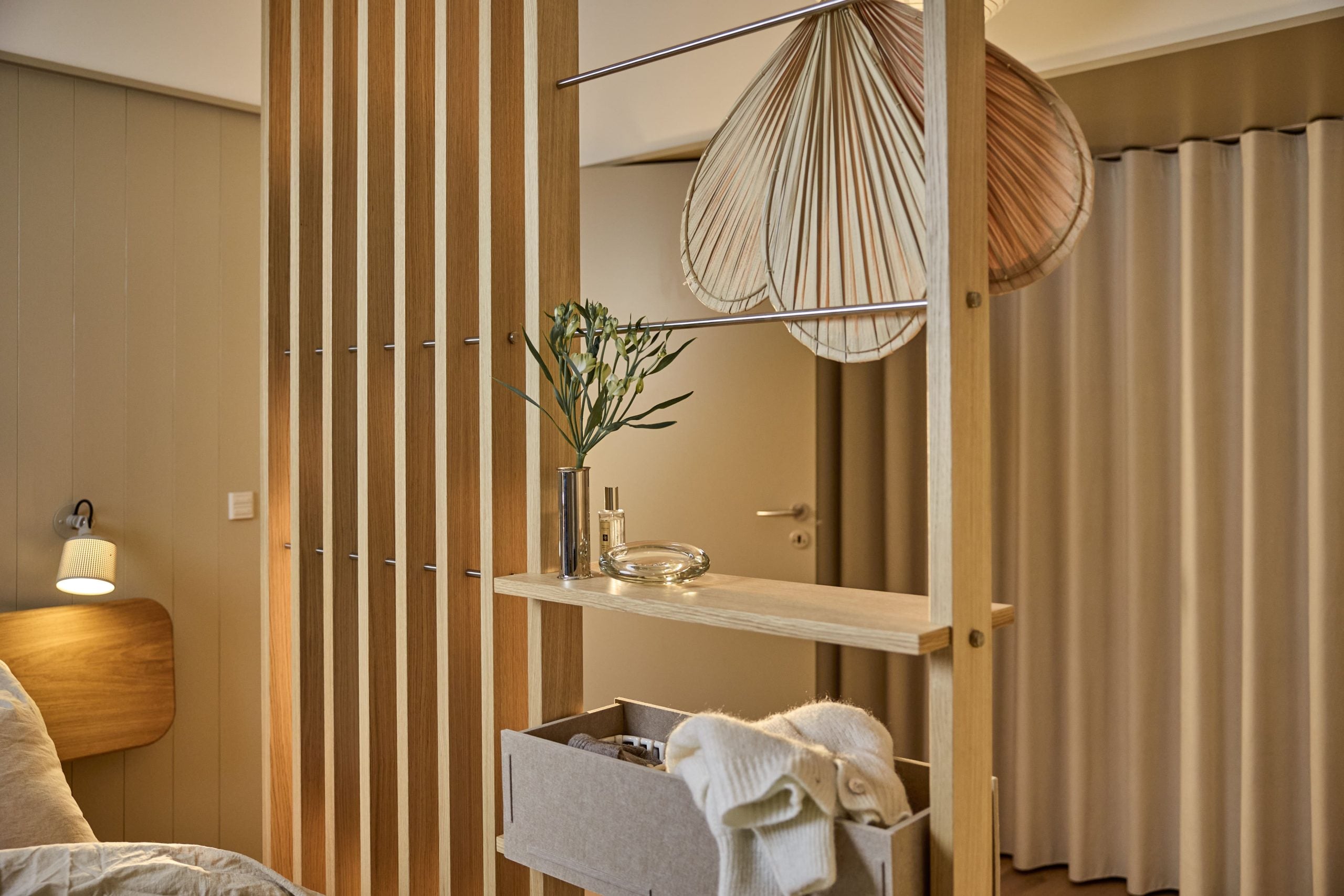 Modern bedroom with a Nordic EMBRACE wooden room divider, shelf holding a plant and towels, glass bottle, and soft lighting for a minimalist atmosphere.