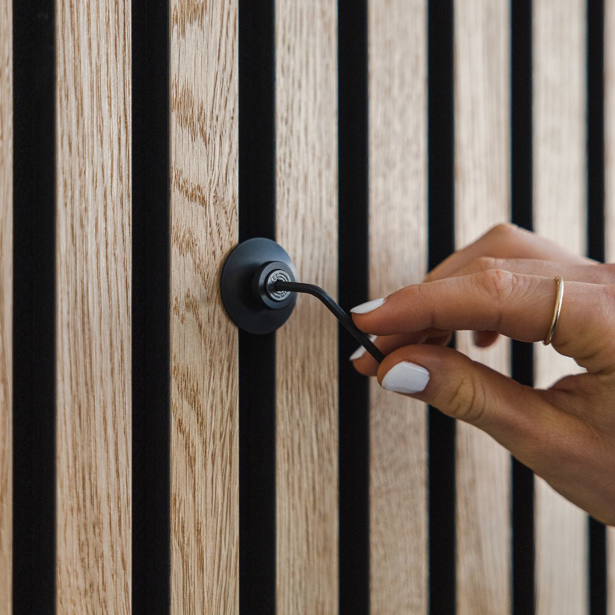 En hand med vitt nagellack vrider på en Dupps | Heavy krok i aluminium black från WoodUpp på en akustisk träpanel vertical Akupanel.