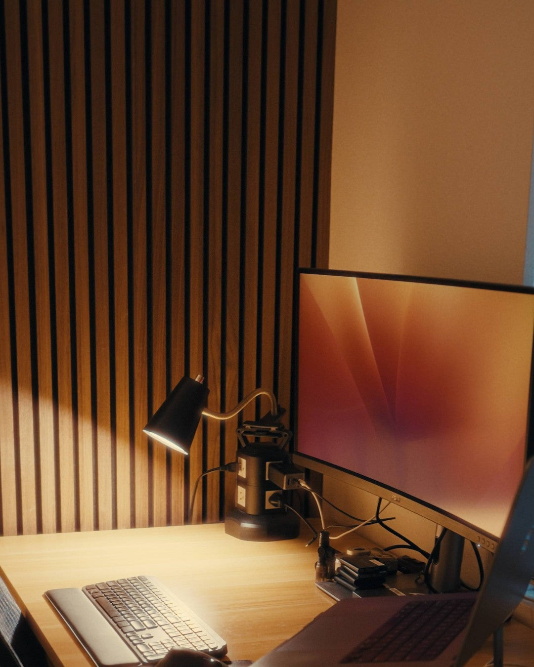 Modern office desk setup with computer monitor, keyboard, and lamp against a wooden slatwall.