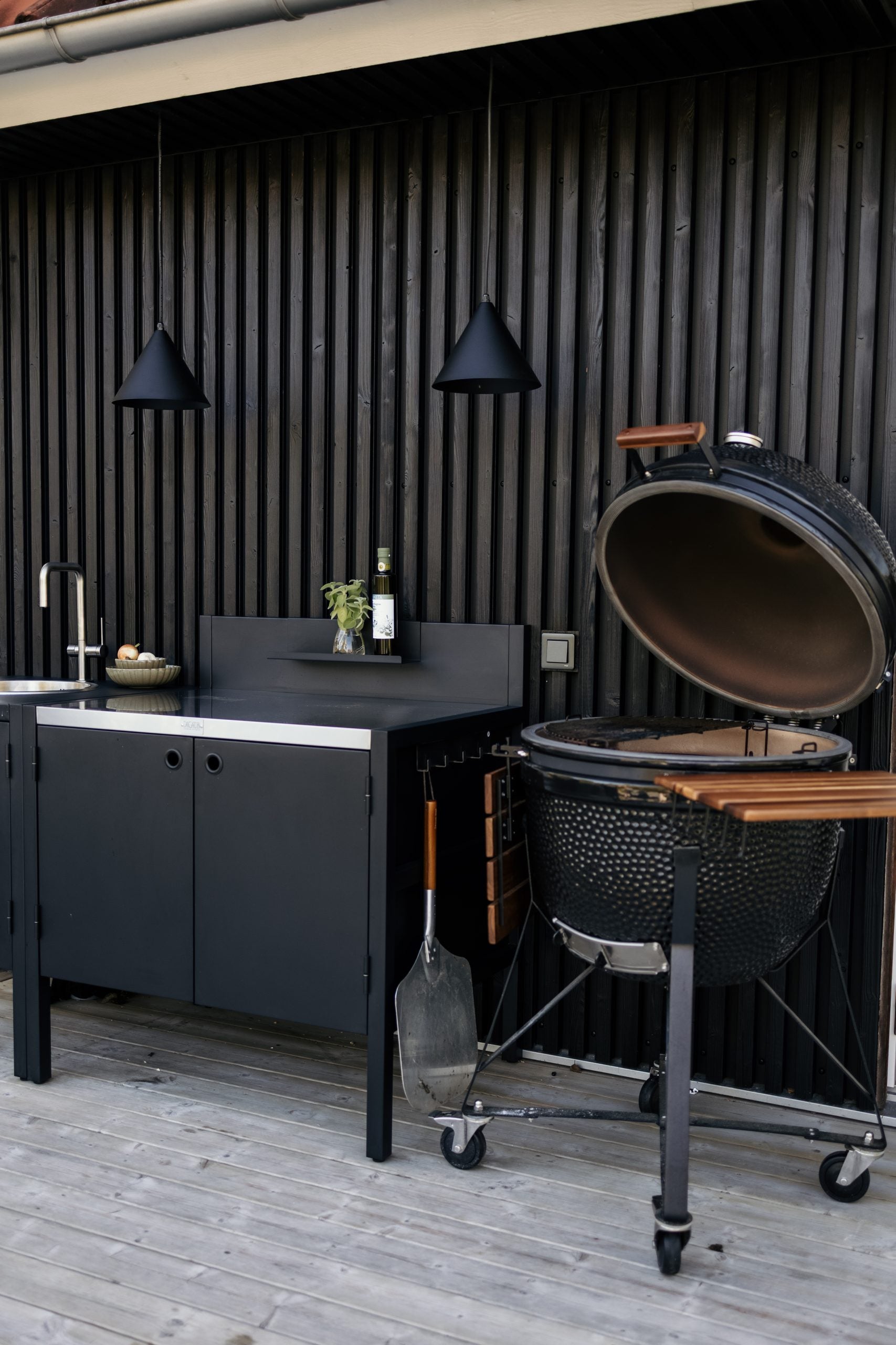Outdoor kitchen with a black sink, cabinets, Kamado-style grill, hanging black lights, and a bottle of wine on a wooden deck with Aluwood facade panels.