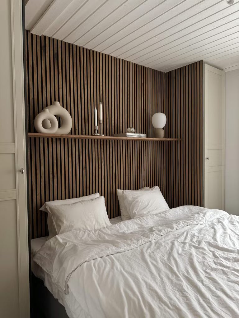 A bed with white bedding is placed against a textured Nordic Akupanel acoustic wood slat accent wall, with minimalist decor and a shelf holding books and plants.
