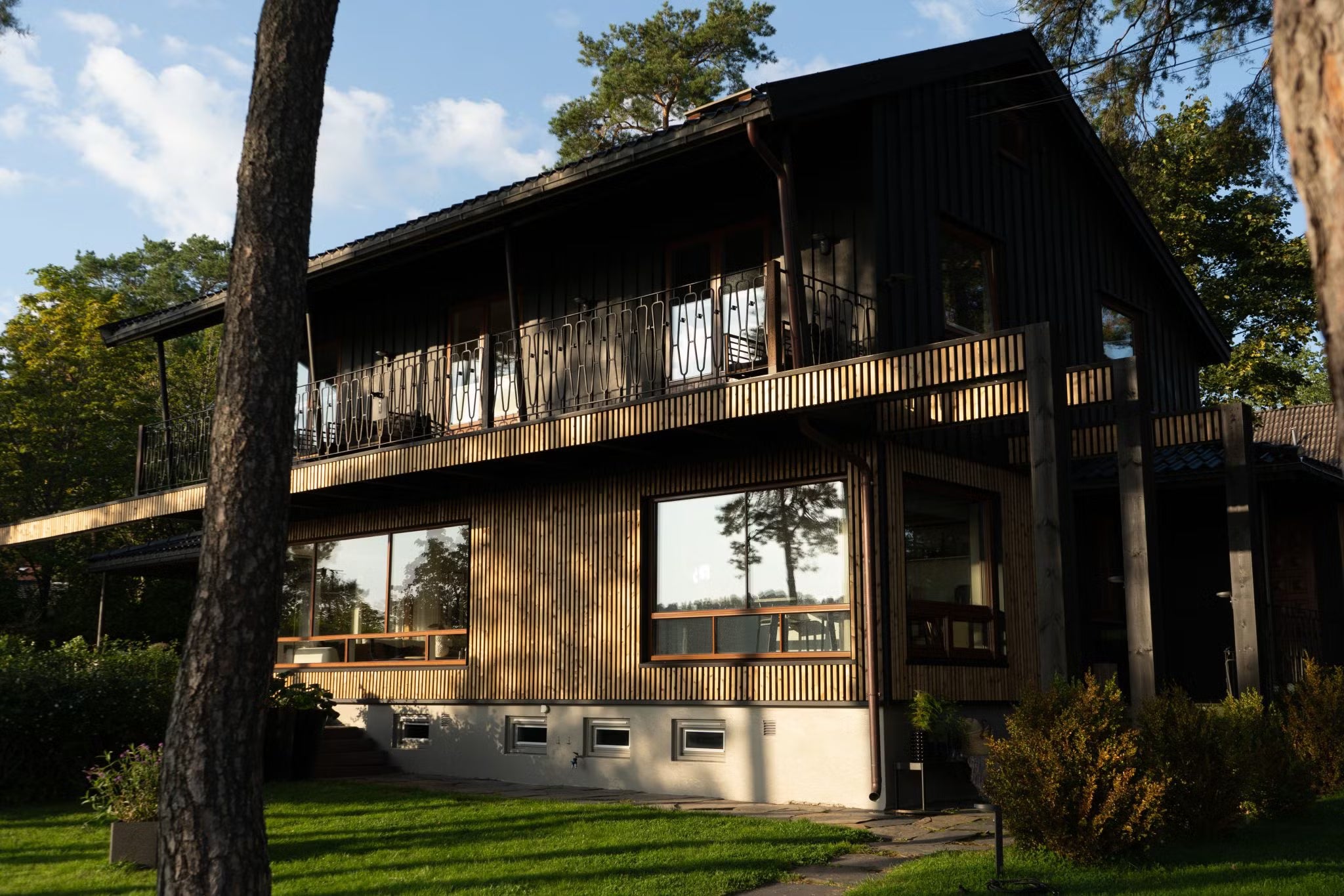 Modern two-story house with large windows and dark wood Aluwood facade panels, surrounded by trees and a green lawn under sunlight.