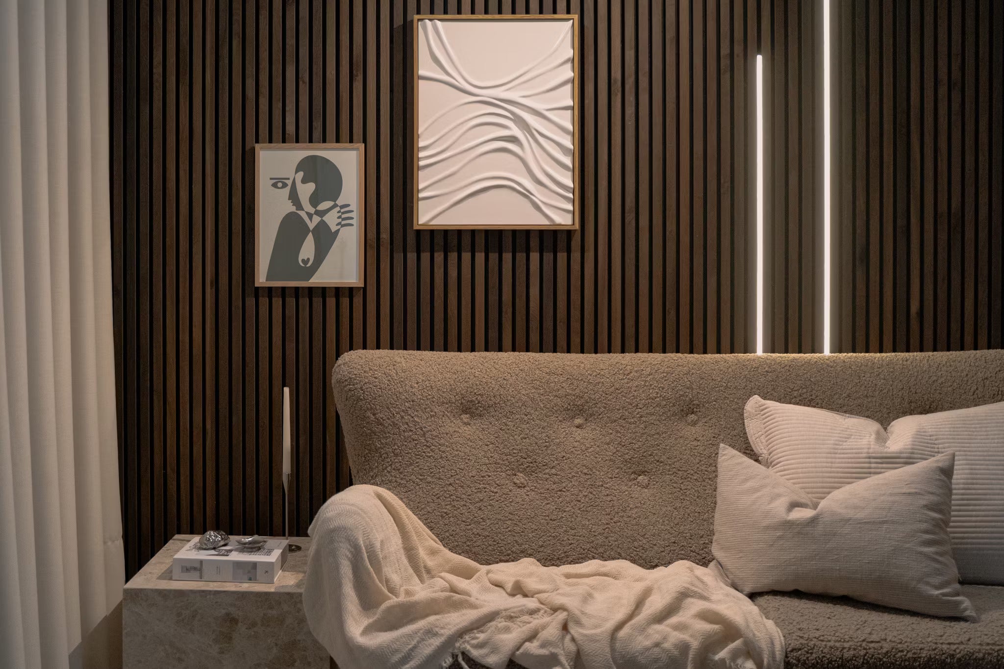 Modernt vardagsrum med beige soffa med textur, vita kuddar, abstrakt konst i akustisk panel och vertikal Akupanel-vägg med träribbor med mjuk belysning.