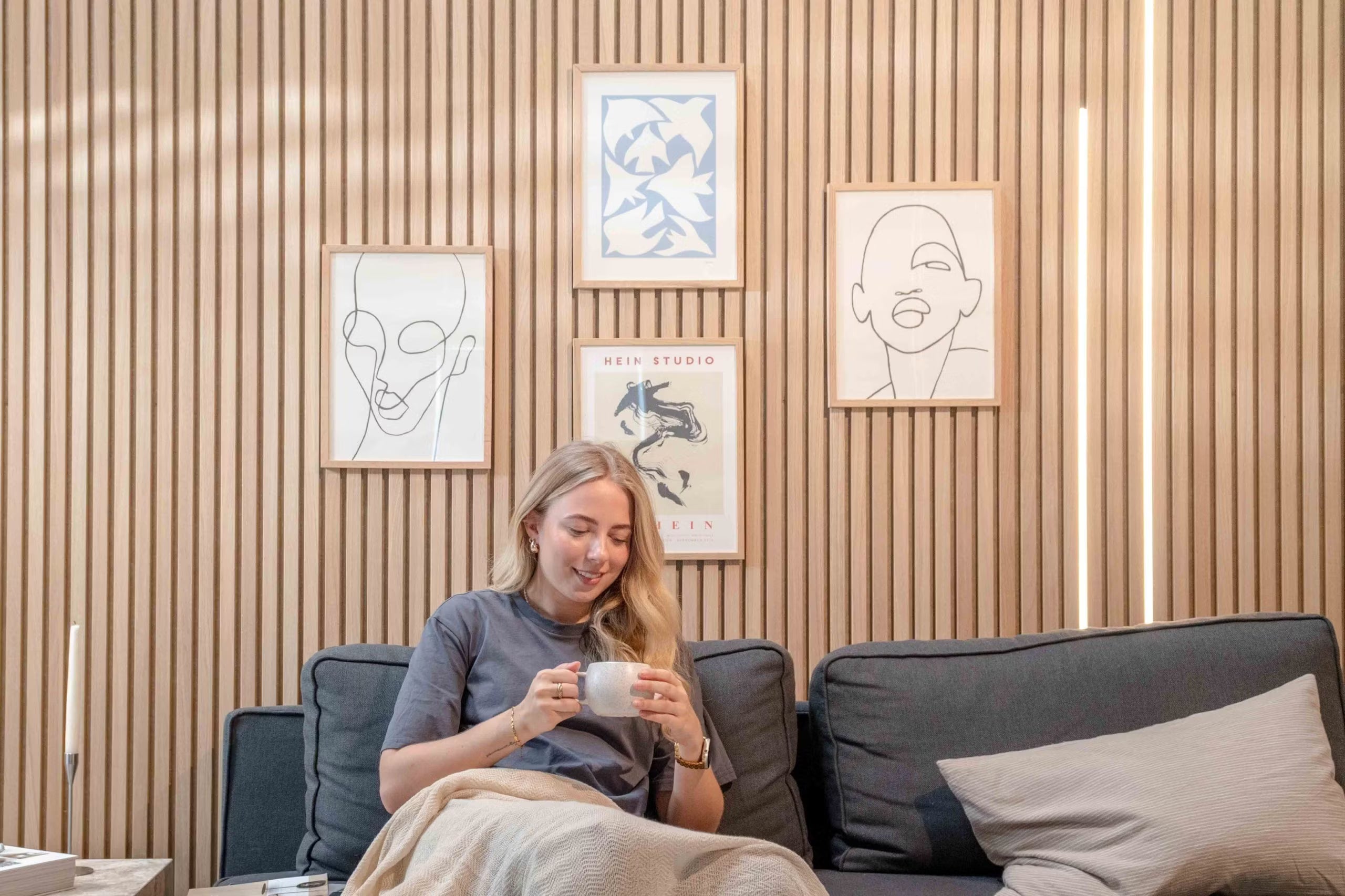 Kvinna som slappnar av i en soffa med en mugg och ler under minimalistisk linjekonst och abstrakt dekor monterad på texturerade nordiska akustiska slatwallpaneler.