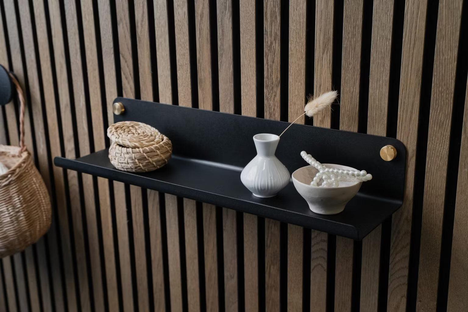 En svart vägghylla med en liten flätad korg, en vit vas och en keramisk skål med pärlor monterad på en nordisk akustikpanel av trä.