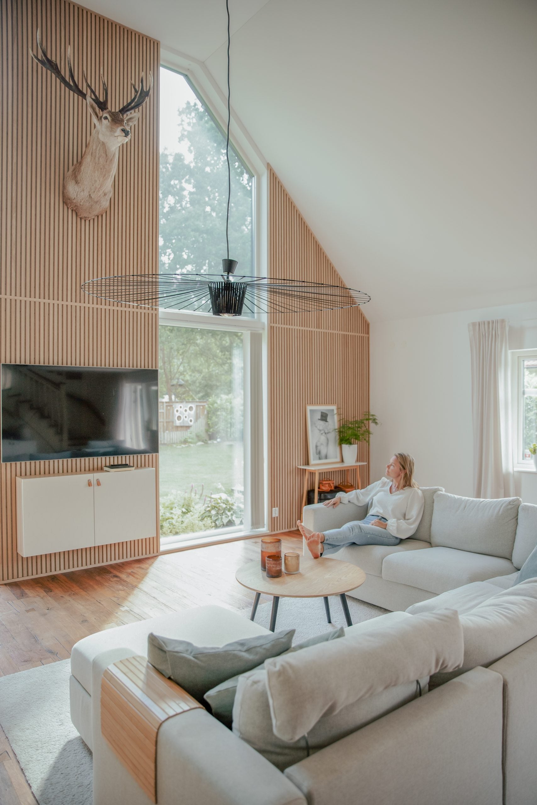 A woman relaxes on a light gray sectional in a bright living room featuring tall windows, Nordic wood accents, and an Akupanel acoustic panel with a mounted deer head.