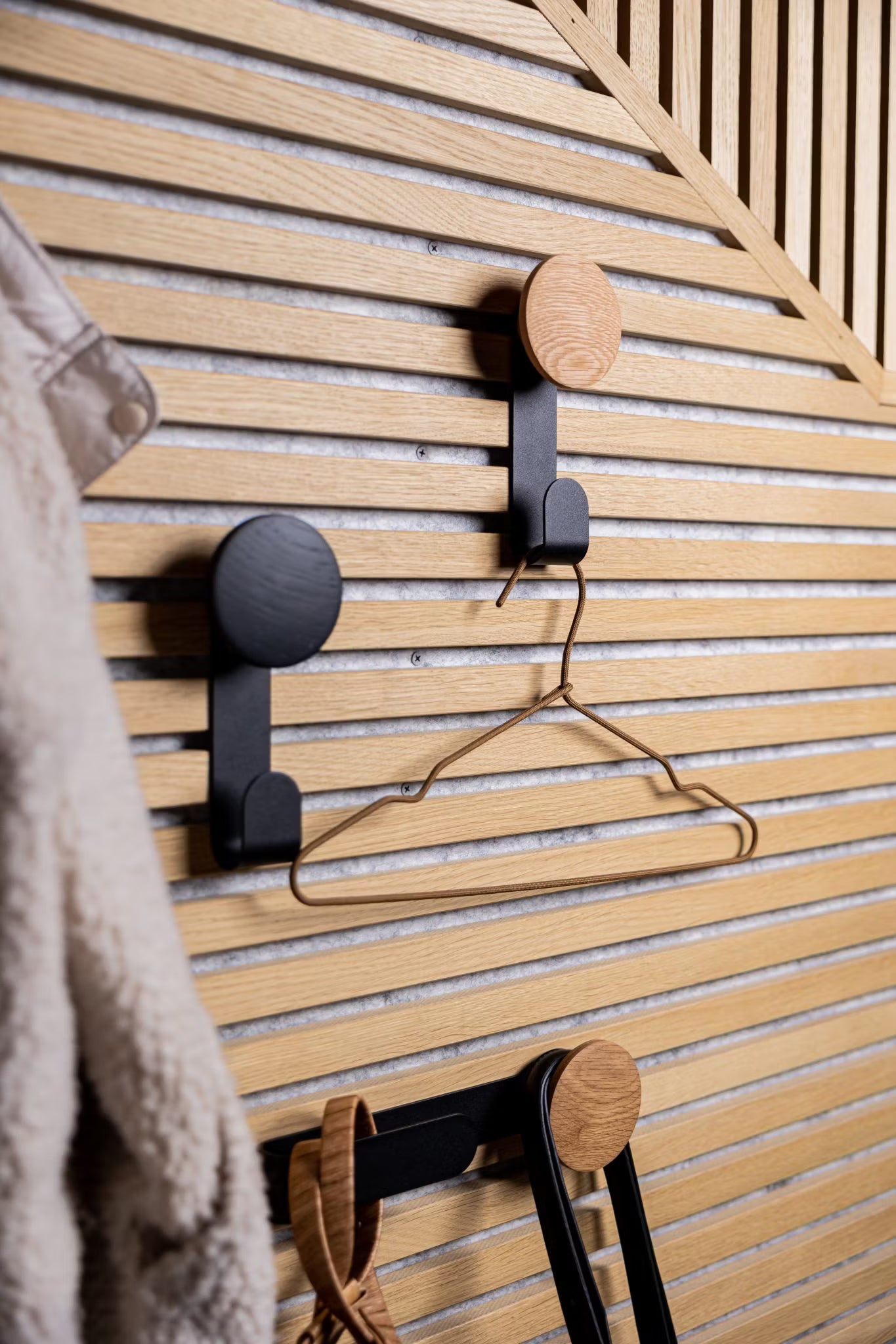 Tre moderna krokar monterade på en Akupanel akustisk träribbvägg i nordisk stil håller en metallgalge och accessoarer, med en luddig kappa delvis synlig till vänster.