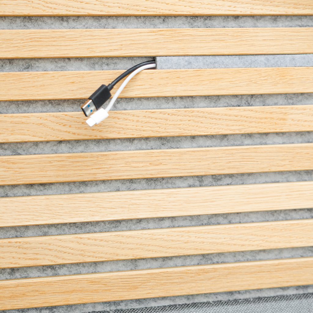 WoodUpp:s eklamellpanel i ljust, naturligt oljad ek med smala horisontella trälameller och grå akustikfilt bakom; en diskret kabelgenomföring visar två kablar som sticker ut — en svart USB-A-kabel och en kort vit laddkabel. Panelen visar träets ådring och ren, modulär design.