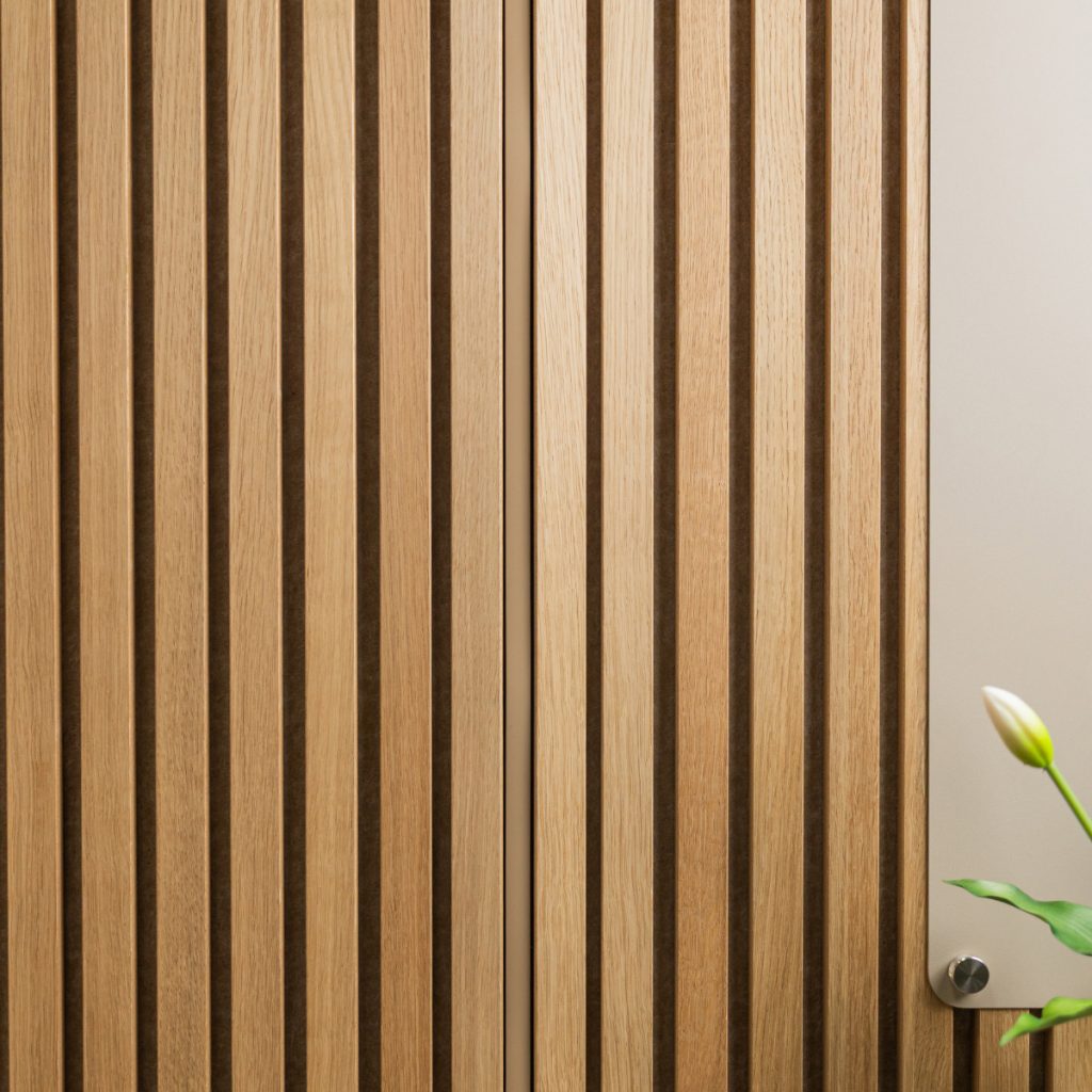 WoodUpp-lamellvägg i ljust trä med jämna lodräta ribbor och mörk bakgrund visar WoodUpps slatpaneler i ek. Till höger syns en ljus monteringsskiva fäst med ett blankt metallfäste och en vit tulpan med gröna blad i bildens nedre hörn med naturlig yta.
