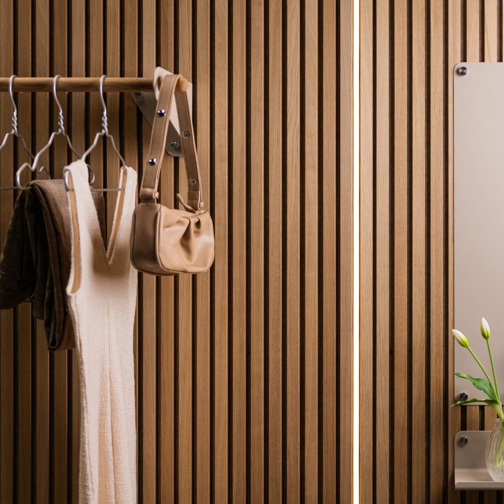 WoodUpp: Vertikal ribbad träpanel i varm ekfinish täcker bakgrunden, med en smal ljuslist mellan skivorna. WoodUpps väggmonterade klädstång med metallfästen bär galgar, en vit stickad klänning, mörkt plagg och en beige axelväska. Till höger en WoodUpp-spegel med liten hylla och vas.