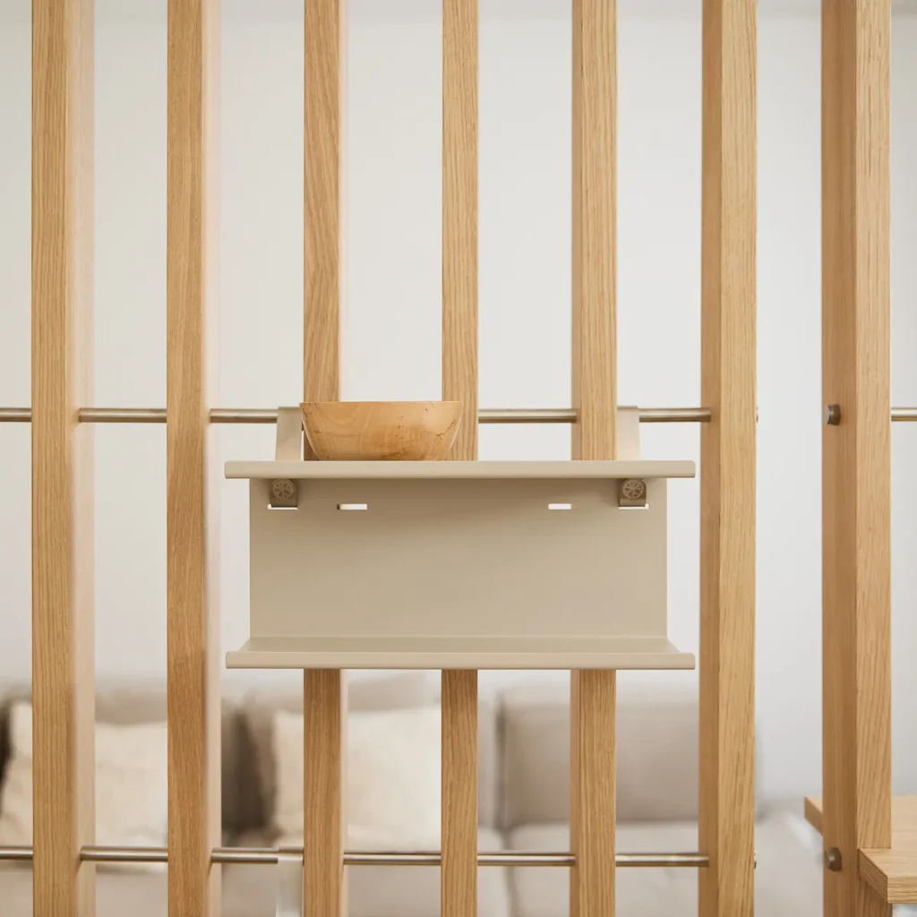 Bild av WoodUpp rumsavdelare i ljus ek med vertikala lameller och metallstänger, med en monterad beige WoodUpp-hylla där en liten träskål står. Enkel, modulär förvaring med synliga fästen och rundade hyllkanter, stilren, neutral palett i mjuka toner och suddad soffa i bakgrunden.