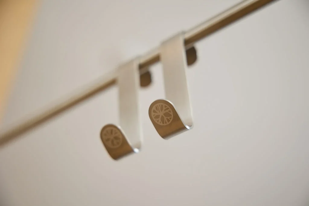 Närbild av två WoodUpp-krokar i borstat rostfritt stål som hänger över en tunn stång. Enkla S-formade upphängningar med präglad rund logotyp på framkanten. Produkterna syns utan textil, visar designen och den släta, mattmetalliska ytan tydligt, perfekt för gardiner eller klädhängare i minimalistiska hem.