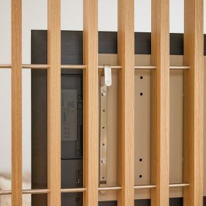 Bild visar WoodUpp:s väggpanel med vertikala eklameller och tunn rostfri stag, monterad bakom en TV. WoodUpp:s dekorativa slatwall i ljus ek, synligt TV-fäste i borstat stål och TV:ns baksida framgår tydligt, inklusive fästöglor och monteringshål. Detaljer som skruvhål, monteringsplatta och diskret hängkrok syns nära lamellerna.