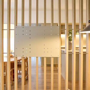 En WoodUpp trälamellvägg i ljus ek med vertikala spjälor och tunna metallstänger, framför vilken en WoodUpp monteringsplatta för TV/fästen sitter monterad — en matt beige perforerad metallplatta med flera fästhål. Stabil, diskret design med slät yta i fyrkantigt format, centralt monterad på lamellerna.