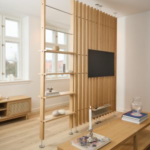 WoodUpp rumsavdelare i ljus ek med vertikala lameller, fästa i tak och golv, med integrerat TV‑fäste och smal flytande hylla. Bredvid står en fristående WoodUpp hyllmodul i ek med öppna plan för prydnader, i ett ljust, skandinaviskt vardagsrum. Diskreta metallstänger och runda fötter ger stabilitet och en modern, luftig känsla.