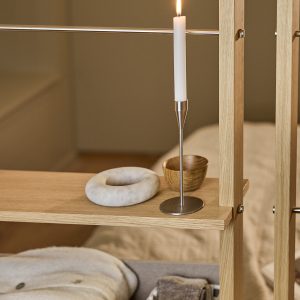 WoodUpp:s öppna hyllsystem i ljus oljad ek med tunna rostfria stålskenor visas här. På hyllplanet står en slank, borstad WoodUpp metalljusstake med vitt kronljus och slimmat fotstöd, intill en rund marmordekoration och en liten handdrejad träskål med varm ådring. Hyllan har diskreta metalldetaljer.