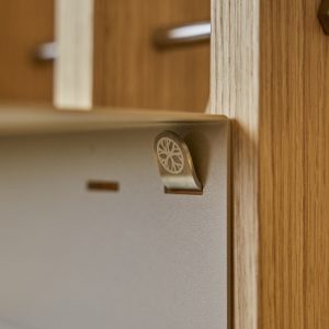 Närbild på WoodUpps metallklämma/knopp fäst i ett ljust ekträstativ; en matt, mörkgrå plåtplatta hänger i klämman. Klämman har ett runt, dekorativt hjulmönster ingraverat. Detaljer som träets ådring, den mjuka, borstad ytan på klämman och avlånga urtag i plåten syns tydligt.